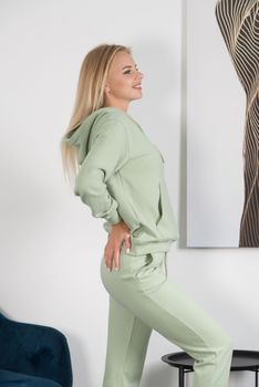 Stylish beautiful young blond woman in a light green tracksuit poses near a white wall in the room. Attractive girl model posing near blue chair.