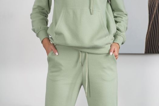 Stylish beautiful young blond woman in a green tracksuit poses near a white wall in the room.