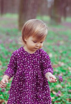blond baby in a colored dress holding flower in a forest glade