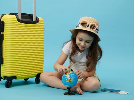 Cute little girl, tourist traveler sitting barefoot near suitcase, choosing a travel destination on the ,blue background