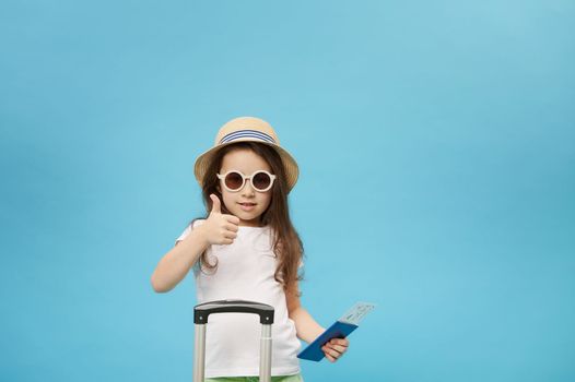 Traveler child girl in sunglasses, holding flight ticket, thumbing up, going for weekend getaway, isolated blue backdrop