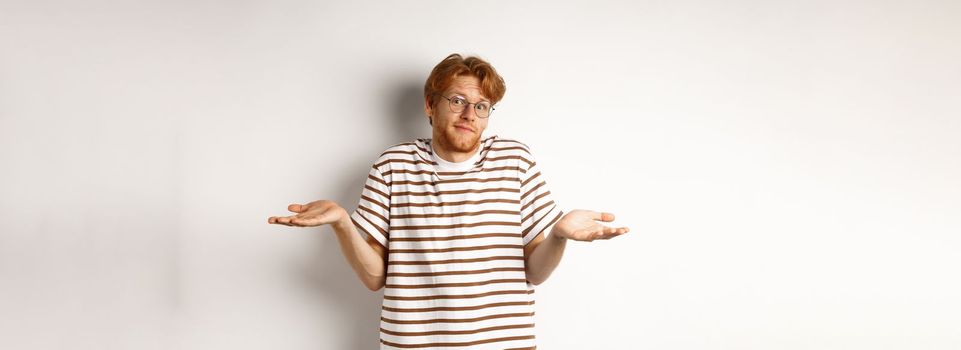 Confused young man with red hair, shrugging shoulders and looking indecisive, standing over white background