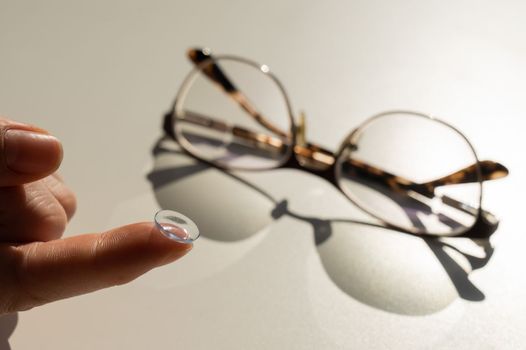 Close-up of a contact lens on a female index finger against the background of glasses on a white table.