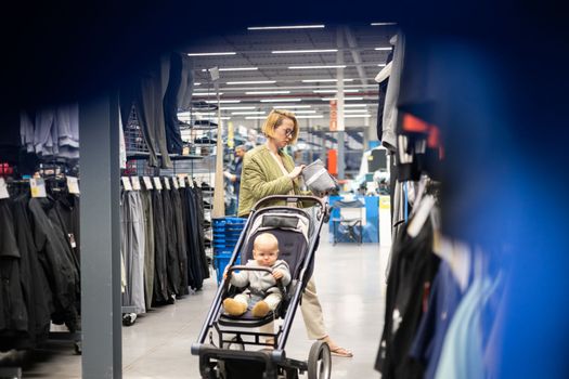 Casualy dressed mother choosing sporty shoes and clothes products in sports department of supermarket store with her infant baby boy child in stroller.
