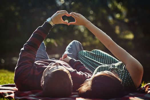 True love is everything. a young couple lying together on the grass and making a heart gesture with their hands.