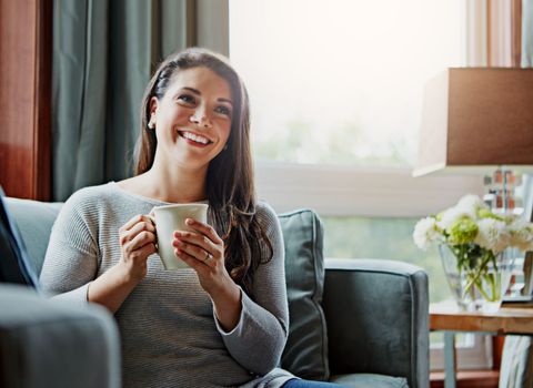Coffee, woman and smile of a relax person at home on a living room sofa relaxing in the morning. Thinking, happy female and lens flare on a lounge house couch with tea feeling happiness with a mug