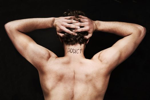 Branded as an addict. Rear view of a naked young man holding his head with the word addict on the back of his neck.