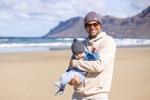 Father enjoying pure nature holding and playing with his infant baby boy sun in on windy sandy beach of Famara, Lanzarote island, Spain. Family travel and parenting concept.