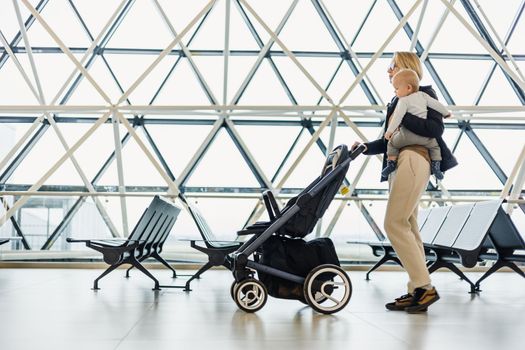 Mother carying his infant baby boy child, pushing stroller at airport departure terminal moving to boarding gates to board an airplane. Family travel with baby concept.