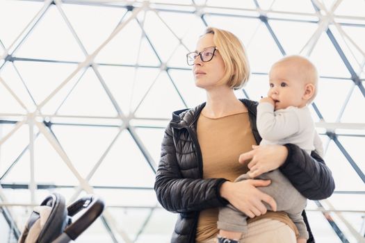 Mother carying his infant baby boy child, pushing stroller at airport departure terminal waiting at boarding gates to board an airplane. Family travel with baby concept.