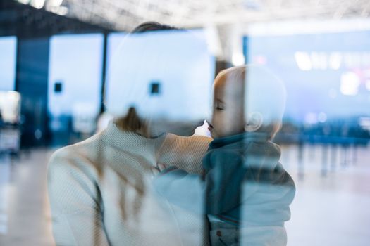Thoughtful young mother kissing and caressing his infant baby boy child while waiting by the window at airport terminal departure gates. Travel with baby concept.