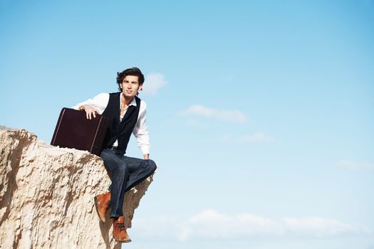 On the edge of the world. Young businessman sitting with his legs dangling over the side of a cliff next to copyspace.