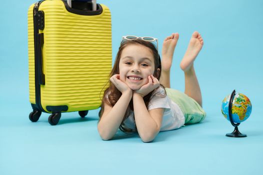 Little girl smiling a toothy smile looking at camera, posing with globe and yellow suitcase, isolated on blue background