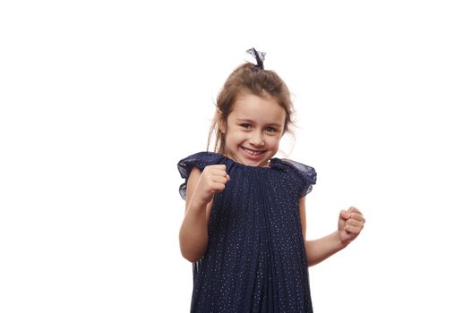 Emotional portrait mischievous little child girl in beautiful elegant blue dress, clenching fists and smiling at camera