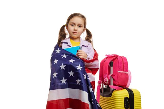 European smart little girl wrapping in U.S.A flag, standing next to suitcase. Travel for study, exchange student concept