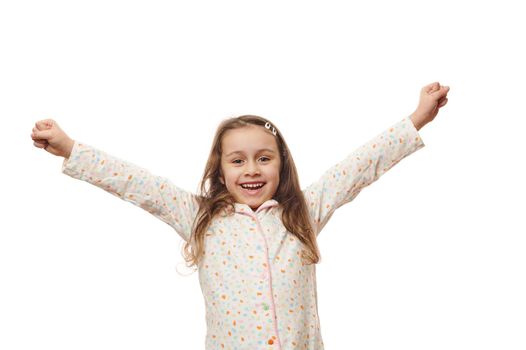 Cheerful baby girl in white pajamas with colorful dots, raising arms up, expressing amazement and happiness, on white