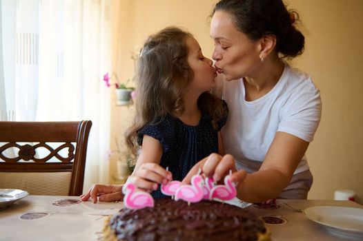 Loving mother kissing her adorable lovely child, daughter while putting candles on birthday cake.