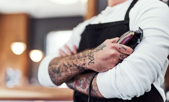 Ill make you look hip and trendy in no time. an unrecognizable tattooed barber posing with his arms folded inside a barbershop.