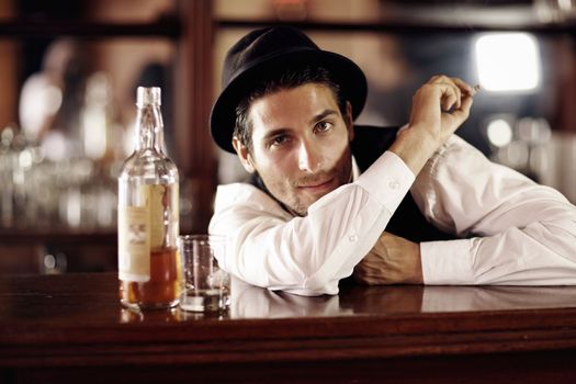 Its been a long day. Portrait of a handsome young bartender smoking with a bottle of whiskey next to him.