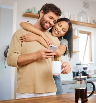 Happy young interracial couple being loving and affectionate at home. Young caucasian man using his smartphone and drinks coffee while his girlfriend embraces him from behind while they look at digit