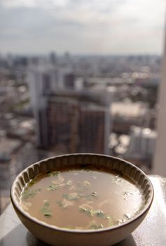Chicken broth with coriander in white bowl over marble table with city view at morning. Selective focus.