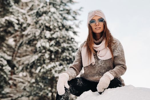 A girl in a sweater and glasses in winter sits on a snow-covered background in the forest