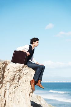 Pondering the meaning of life. Pensive young man sitting with his briefcase on the edge of a cliff overlooking the ocean.