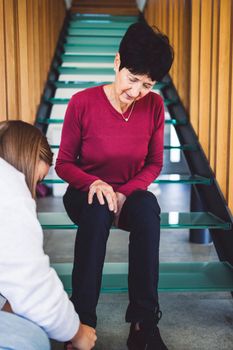 Vertical photo of senior woman sitting on the glass stairs while unrecognizable nurse helps her to put on shoes