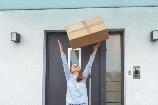 Waist up caucasian woman throwing an exciting package in the air, standing outside her front door