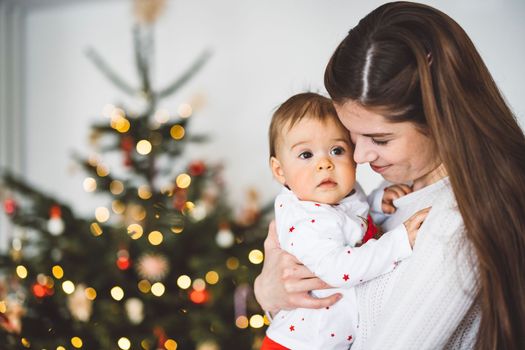 Loving mother holding her baby girl, standing in front of the Christmas tree