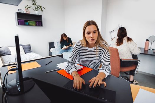 Female student doing homework on her laptop while her roommates are studying in the back
