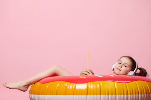Lovely baby girl listens to music in headphones, looking at camera, holding a coconut drink, lying on a donut swim ring
