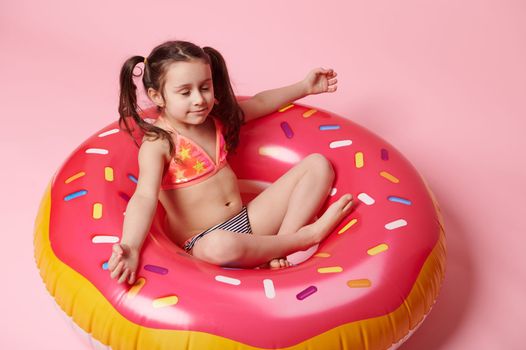Adorable little child girl meditating in lotus position on a pink inflatable swim ring donut, over pink color background