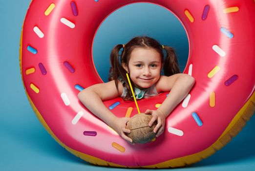 Cute baby girl with coconut water inside pink inflatable swim ring, smiles looking at camera, isolated blue background