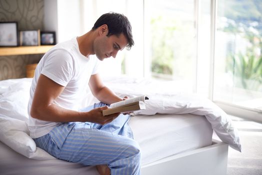 Hes quite the bookworm. a handsome young man reading a book on the edge of his bed.