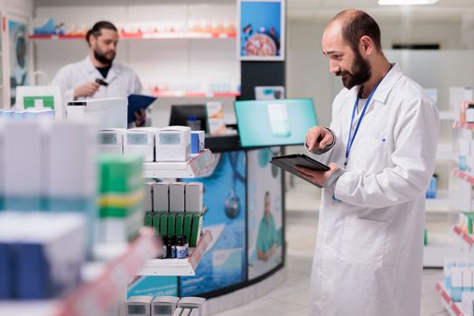 Drugstore worker looking at pills packages typing medicaments name on tablet computer during inventory