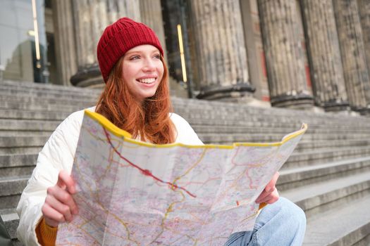 Portrait of young redhead woman, tourist sits with paper map and looks for a route to tourism attraction, rests on stairs outdoors