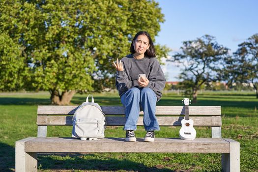 Girl with sad face, sitting on bench with smartphone and ukulele, looking upset and disappointed, being alone in park