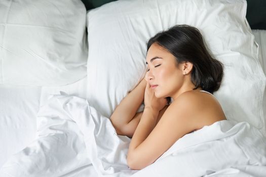 Top view image of asian woman sleeping alone in king size bed on white pillows. Young girl lying in her bedroom with eyes closed, morning sunlight shines in room