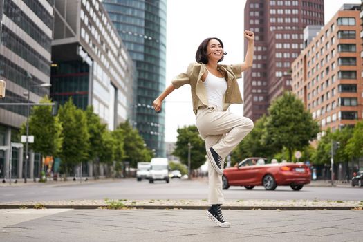 Portrait of asian happy girl jumping and dancing in city centre, posing on streets, express joy and excitement