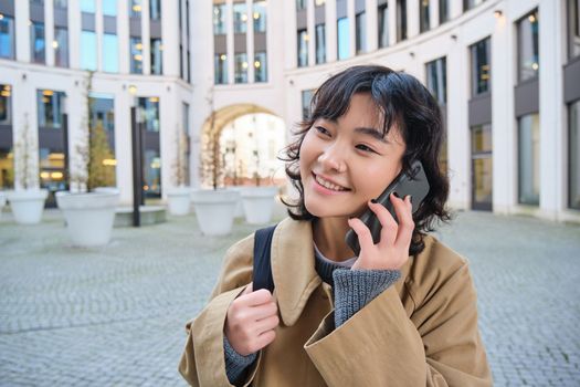 Cellular technology. Young korean woman talks on mobile phone, makes a phone call on her way home, walks down street, city centre, has telephone conversation