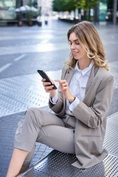 Vertical portrait of businesswoman in suit using her phone, relaxing outdoors, sitting on bench