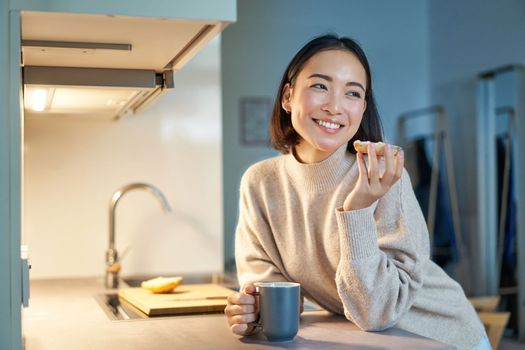 Beautiful young asian woman eating toast on her kitchen, drinking coffee and smiling