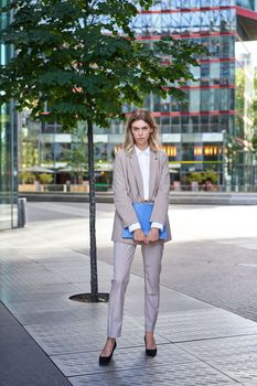 Portrait of young saleswoman in beige business suit, holding blue folder with work documents, standing outdoors on street of city center