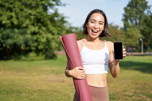 Smiling asian fitness girl with rubber yoga mat, shows her smartphone screen, recommends workout application, stands on lawn in park.