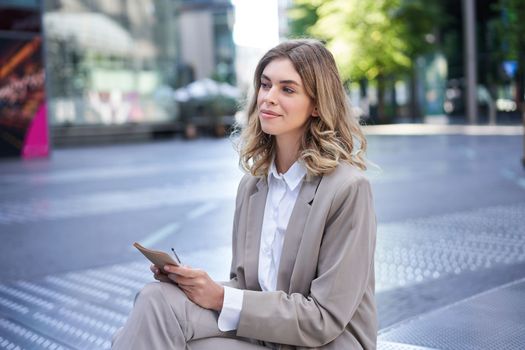 Young businesswoman preparing for speech, interview in company, writing down notes while sitting outside in city centre