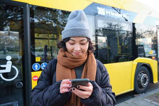 Image of girl student waiting for public transport, checks schedule on smartphone app, stands near city bus