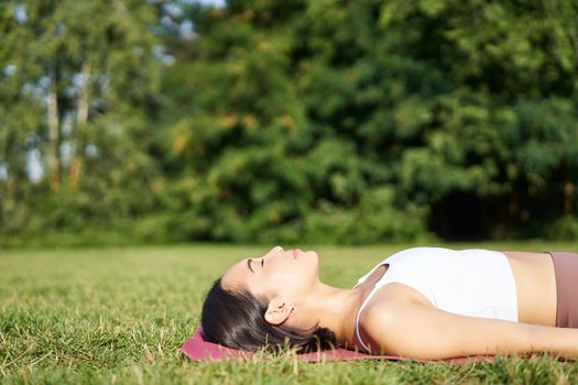 Young fitness girl lying on sport mat on lawn, breathing and meditating in park in sportswear.