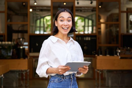 Smiling beautiful waitress, businesswoman with digital tablet, laughing and looking happy, standing in front of cafe or restaurant