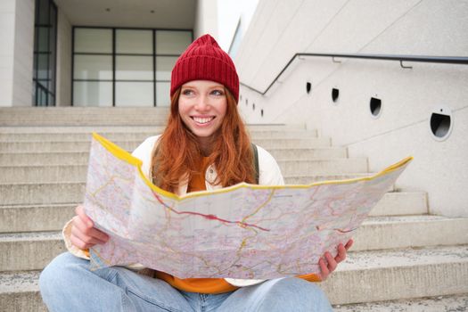 Beautiful girl tourist sits on stairs with city map, plans her journey, looks for direction while travelling around town, searches route for sightseeing